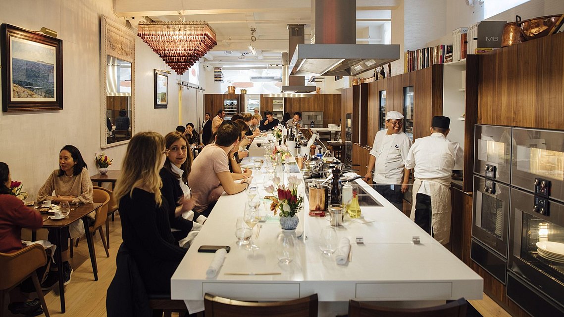 Gäste sitzen am Tresen des Restaurants und sehen zu wie die Köche die Speisen zubereiten.